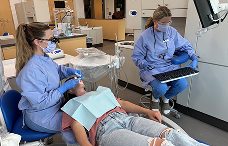 two students in full scrubs and ppe clean a patient's teeth in a dental office