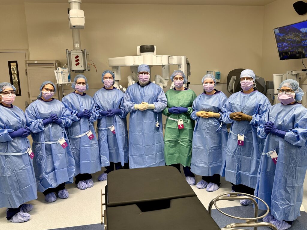 A group of surgical technology students stand in a circle in blue gowns with their hands crossed, wearing blue gowns except for one person in a green gown
